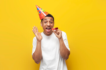 african american guy in festive hat celebrates his birthday and rejoices on yellow isolated background