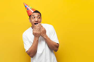 shocked african american guy in party hat celebrates birthday and shows surprise on yellow isolated...