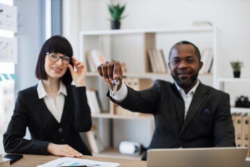 Smiling real estate agents in formal attire holding house keys while sitting at office desk in comfortable workplace. Multiracial man and caucasian woman arranging renting of modern building.