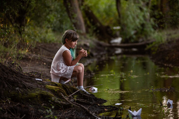 Teenage girl with green hair in dress, stands on the river bank, launches white paper origami boat