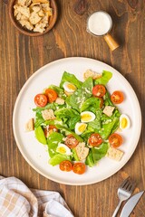 Caesar salad with romaine lettuce, cherry tomatoes, boiled quail eggs and croutons on a light ceramic plate on a wooden background. American cuisine