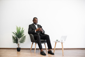 Successful african american businessman in stylish formal suit sitting relaxed in comfy chair and typing on modern smartphone. Cup of coffee and wireless laptop placed on small table near.