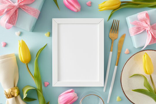 Mother's Day concept. Top view photo of vertical photo frame gift boxes plate cutlery knife fork fabric napkin yellow pink tulips and colorful hearts on pastel blue background with blank space