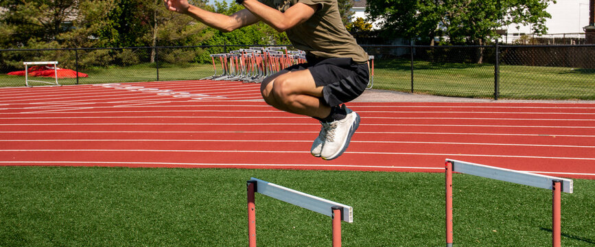 Male athlete in shorts jumping over track hurdle on a turf field - Powered by Adobe