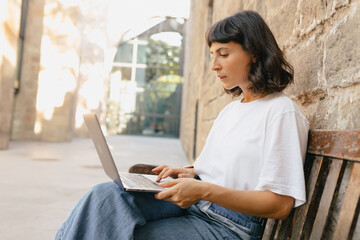 Cute stylish young lady sitting with laptop outdoors and smiling. Pretty brunette girl, wearing light clothes, looking into screen laptop and working