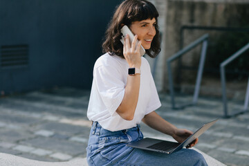 Pretty young caucasian girl with gadgets smiling looking aside spending time outdoors. Brunette wears casual clothes. Concept lifestyle, technology in modern society