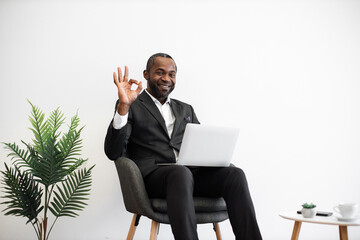 Diligent african employee in formal wear smiling sincerely at camera while typing on wireless laptop at office with modern minimalist design showing sign ok. People, work and technology concept.