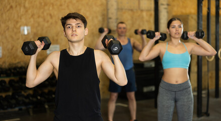 Young dedicated man doing exercises with dumbbells near other people in gym