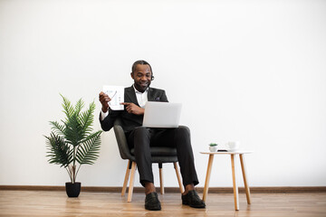 Dedicated african american office worker using headset and laptop for video conference at office. Emotional young man in suit showing graphs while presenting financial report for employer online.