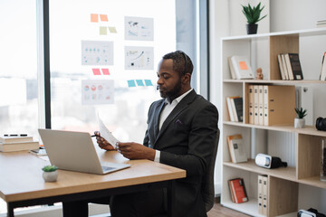 Formally dressed male entrepreneur sitting at workspace and having online meeting with colleagues on laptop. African american man in headset examining some documents holding in hands.