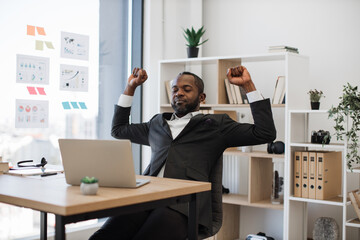 Sleepy and tired african american man in formal wear yawning and stretching arms after hard working hours on wireless laptop at office. Concept of people, technology and overwork.