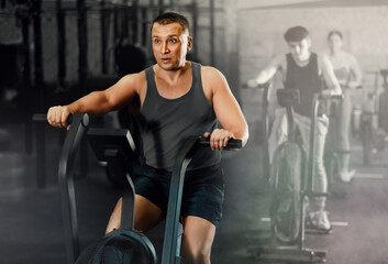 Naklejka na ściany i meble Dedicated man training on exercise bike near other people in gym
