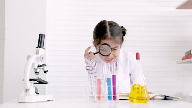 Scientist elementary girl wearing gown suit using magnifying glass to look at chemical test tube in chemistry classroom, Young science kid studying an experiment in laboratory,