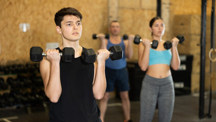 Sportive strong young male in activewear lifting dumbbells during group workout class in gym indoors. Functional training concept