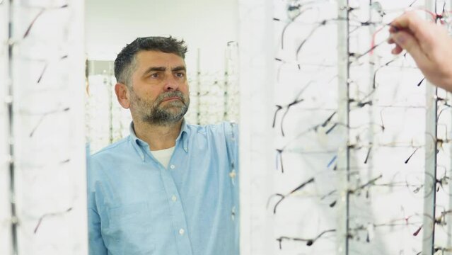 Bearded senior man choosing new glasses at optical shop, looking at mirror