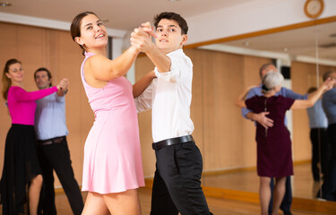 Fototapeta premium Couple of smiling young guy and girl enjoying slow foxtrot in dance studio. Amateur ballroom dancing concept