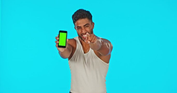 Face, green screen and man with phone pointing in studio isolated on a blue background. Cellphone, thumbs up and happy Indian male flex bicep muscle with advertising, mock up and product placement.