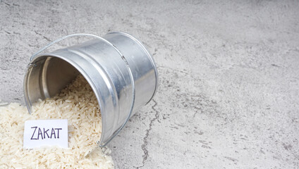 Zakat words with pile of white rice in metal bucket againts cement textured background.