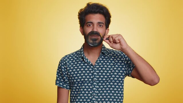 Indian man holding magnifier glass on healthy white teeth, looking at camera with happy expression, showing funny silly face smiling mouth. Handsome hindu guy isolated on yellow studio background