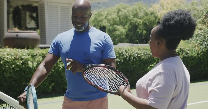 Happy senior african american couple with tennis rackets talking at tennis court in slow motion