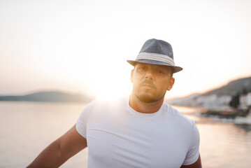 A fit and attractive man on vacation at a hotel near a sandy beach. He is wearing a stylish hat and enjoying the warm sun and crystal-clear sea. The scenery is serene and idyllic, portraying a sense