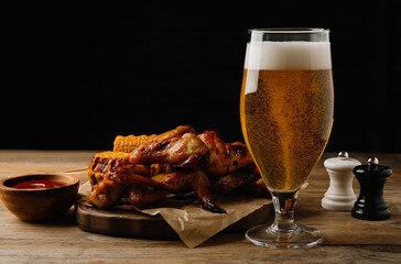 Glass of beer, delicious baked chicken wings, grilled corn and sauce on wooden table against black background