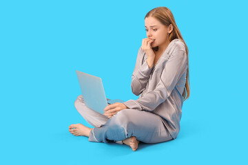 Fototapeta premium Young woman in pajamas with laptop biting nails on blue background