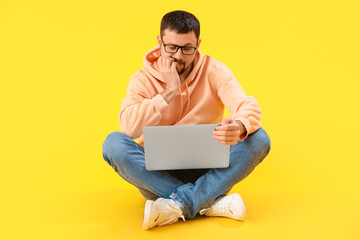 Handsome man with laptop biting nails on yellow background