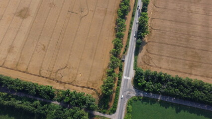 Automobile asphalt road with cars driving between agricultural fields with yellow ripened wheat and different green agricultural crops in summer. Aerial drone view. Agrarian crop panoramic landscape