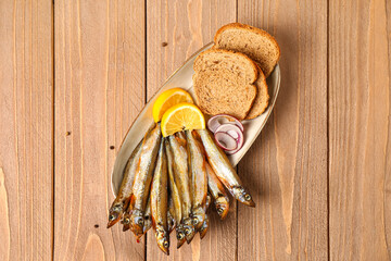 Plate with delicious smoked capelin, lemon and bread slices on wooden background