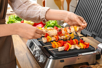Woman cooking tasty chicken skewers and vegetables on modern electric grill at table in kitchen, closeup