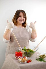 Obraz na płótnie Canvas Cheerful female chef is sitting at the table with bunch of vegetable, showing prepared meal and ok sign.Female chef showing prepared meal and ok sign High quality photo