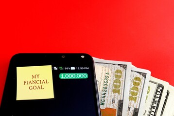 Cash money, smart phone on red background with text note written MY FINANCIAL GOAL, concept of money management , goal setting to save and invest to get rich and be a millionaire