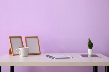 Workplace with tablet computer, notebook and blank frames near lilac wall