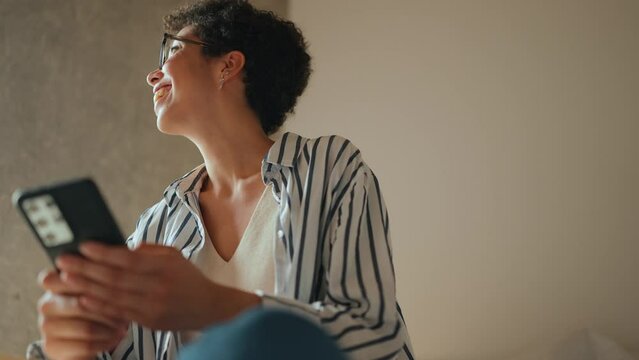 Laughing Hispanic curly haired woman texting on mobile on the bed at home
