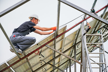 Man engineer installing photovoltaic solar panel system under white sky. Male workers in safety construction helmets mounting solar modules, panels and support structures of photovoltaic solar array.