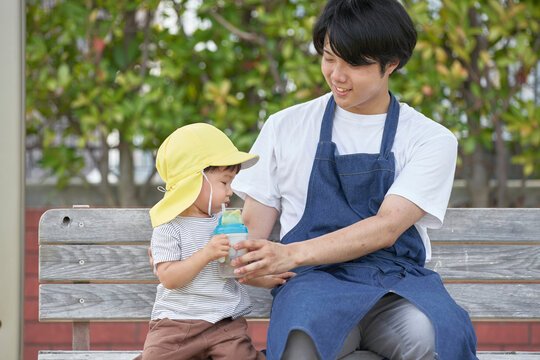 ベンチに座って子供にお茶を飲ませる男性の保育士