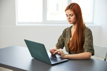 red-haired woman typing in a laptop sitting at a table in the office near the window working full time