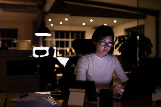 Asian businesswoman wearing glasses, sitting at desk, using laptop working late at office