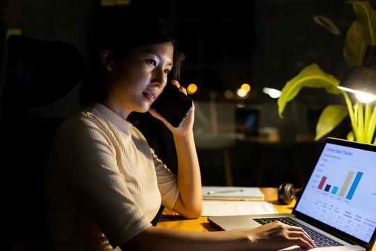 Happy asian businesswoman at desk, using laptop and talking on smartphone, working late at office