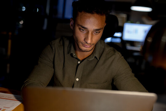 Biracial businessman sitting at desk, using laptop and working late at office