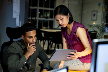 Diverse business people sitting at desk, using tablet and discussing, working late at office