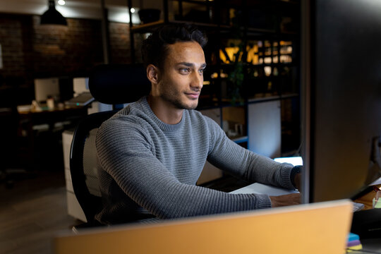 Happy biracial businessman sitting at desk, using computer, working late at office