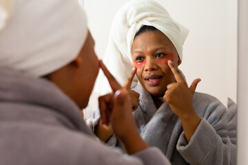 Obraz na płótnie Canvas Happy plus size african american woman wearing robe, applying eye masks, looking in bathroom mirror