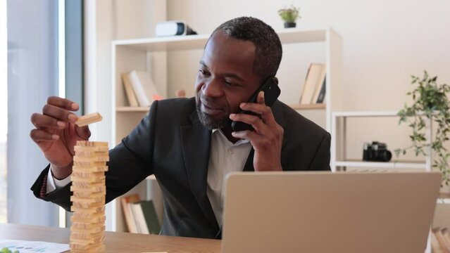 Happy multicultural man in business suit playing jenga while talking on smartphone at office desk with laptop. Cheerful entrepreneur reducing stress at work by enjoying board game during conversation.
