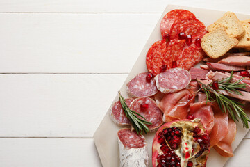 Board with assortment of tasty deli meats on white wooden background, closeup