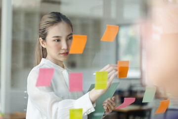 Asian woman working on project plan using sticky papers notes on glass wall, people meeting to share idea, Business design planning concepts.