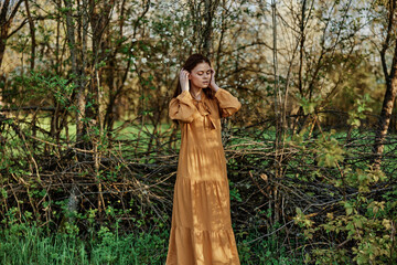 a beautiful woman walks in the shade near the trees, dressed in a long orange dress, enjoying the weather and the weekend, straightening her hair with her hands. The theme of privacy with nature