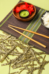 Bamboo mat with ginger, wasabi and cut nori sheets on green background, closeup