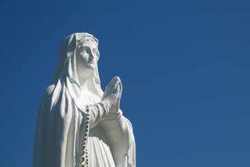 Fototapeta premium White antique statue of Virgin Mary praying against blue sky. Copy space.